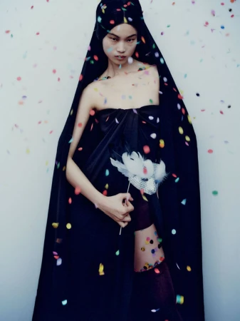 Model in black hooded cloak holding feathered mask amid falling confetti for Mixte