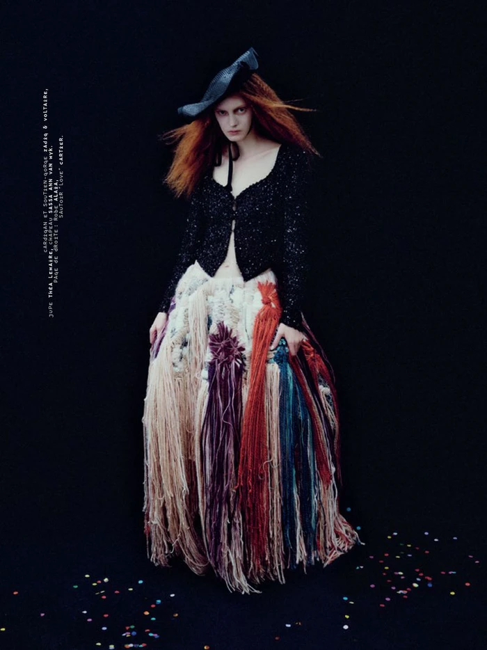 Model with red hair wearing a black cardigan and textured fringed skirt against black