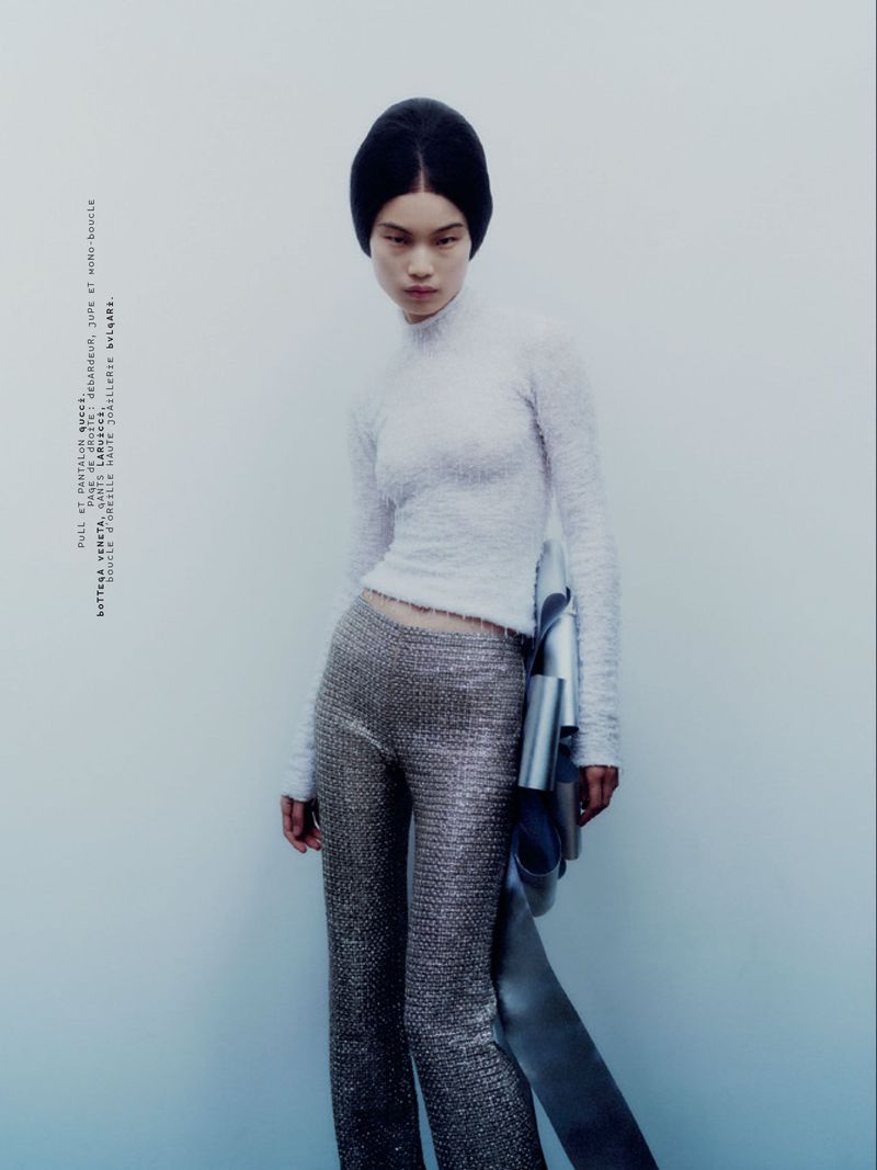 Model in textured white top and metallic trousers holding a silver garment against a pale seamless studio background.