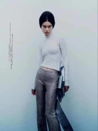 Model in textured white top and metallic trousers holding a silver garment against a pale seamless studio background.