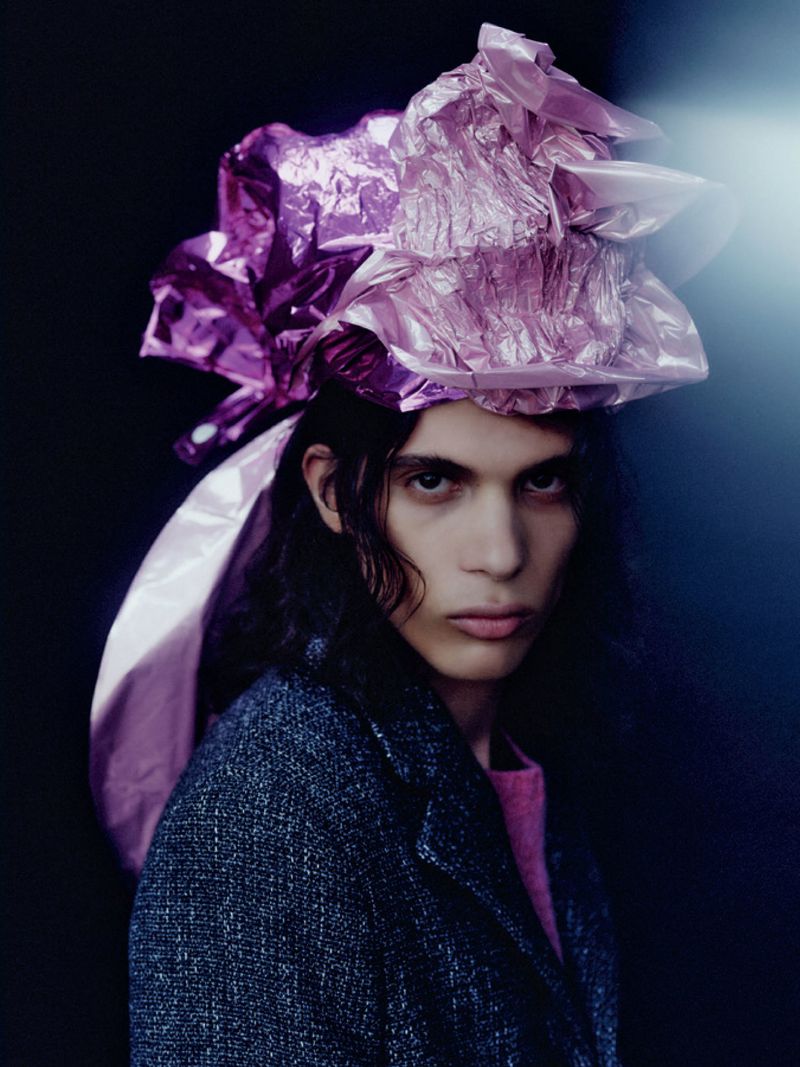 Portrait of a model wearing a crumpled metallic pink wrap on their head and a dark textured jacket against a dark background.