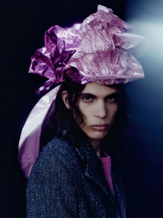 Portrait of a model wearing a crumpled metallic pink wrap on their head and a dark textured jacket against a dark background.