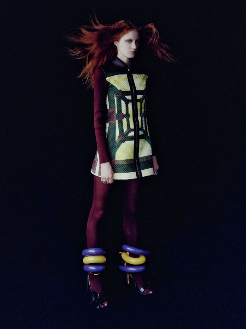 Model with red hair wearing a patterned dress and dark tights with large colorful ankle rings