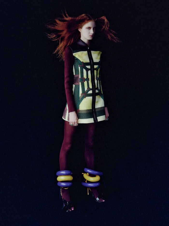 Model with red hair wearing a patterned dress and dark tights with large colorful ankle rings