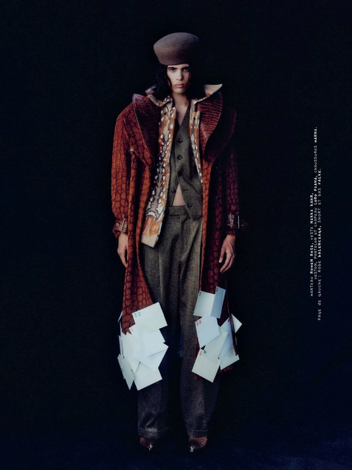 Model standing against a dark background wearing a textured brown coat adorned with hanging cards and a cap for Mixte.