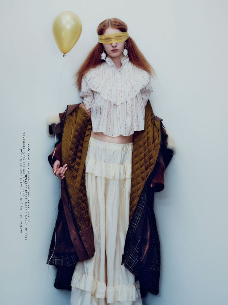 Model with blindfold holding yellow balloon wearing ruffled blouse and coat against plain background