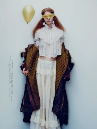 Model with blindfold holding yellow balloon wearing ruffled blouse and coat against plain background