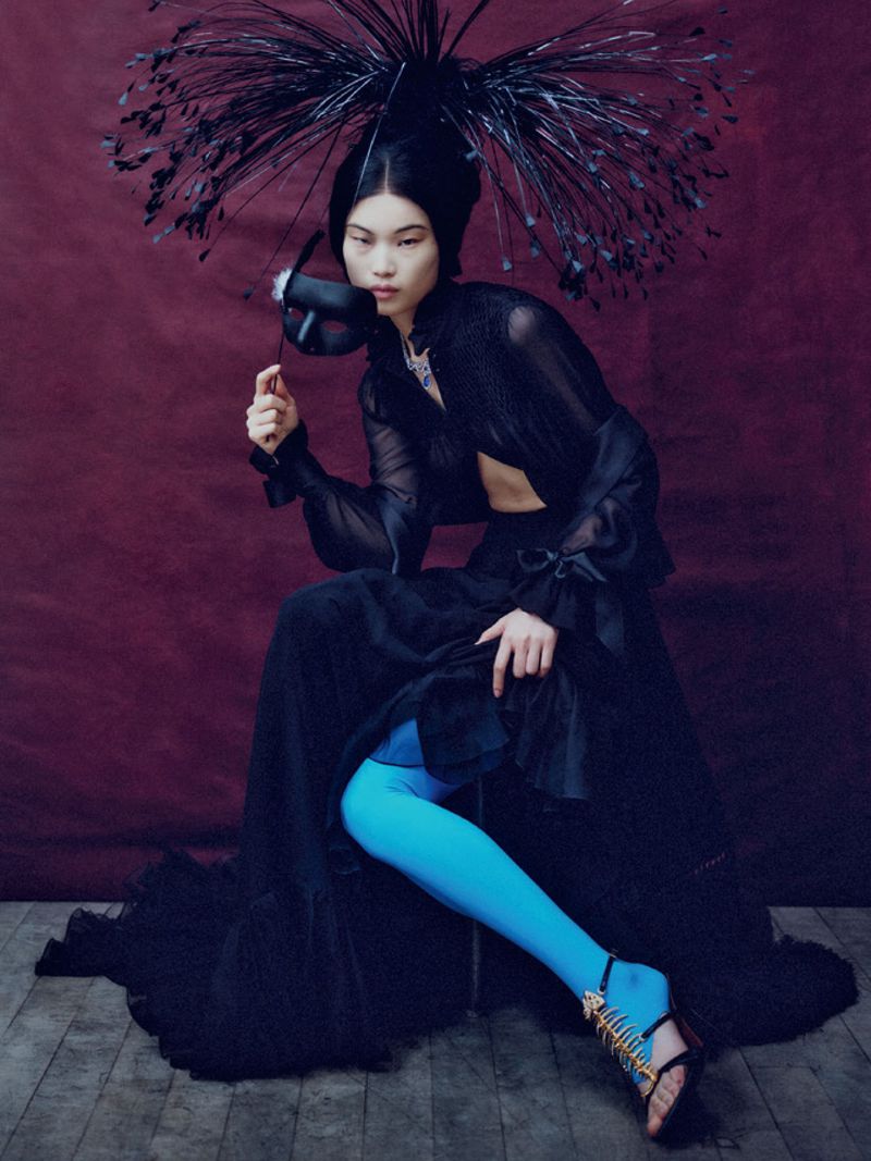Model in black sheer blouse and voluminous skirt holding a mask, wearing bright blue tights and a spiky feathery headdress.