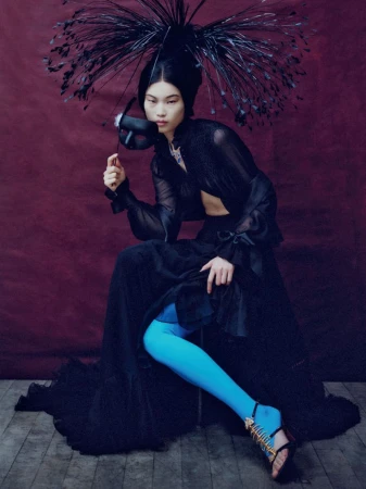 Model in black sheer blouse and voluminous skirt holding a mask, wearing bright blue tights and a spiky feathery headdress.