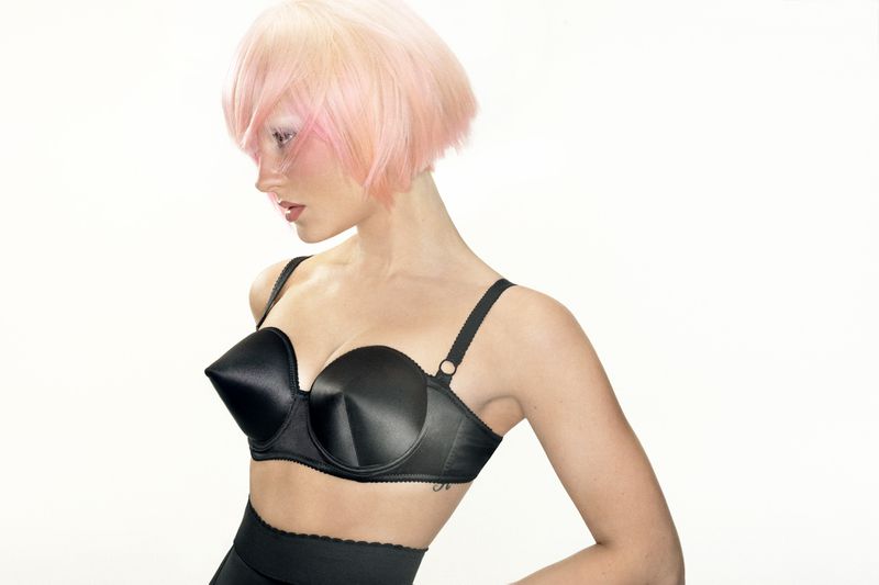 Woman with pink bob hair wearing a black cone bra against a white background for Allure Magazine.
