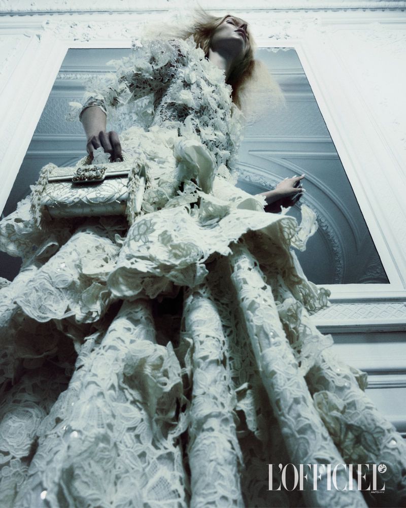 Model wearing a white lace haute couture gown and holding a matching embroidered handbag against an architectural wall.