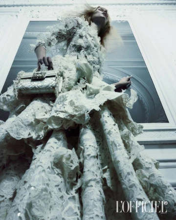Model wearing a white lace haute couture gown and holding a matching embroidered handbag against an architectural wall.