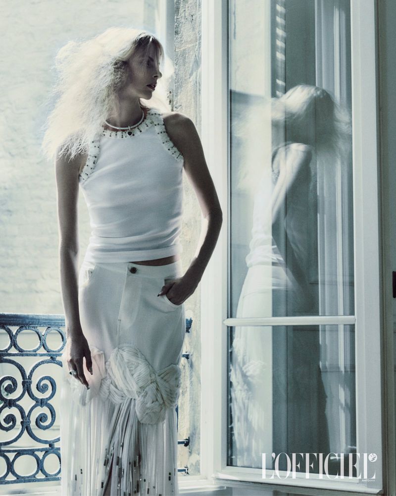 Model Elza Matiz in a white tank top and skirt standing near a large window for L'Officiel Italia