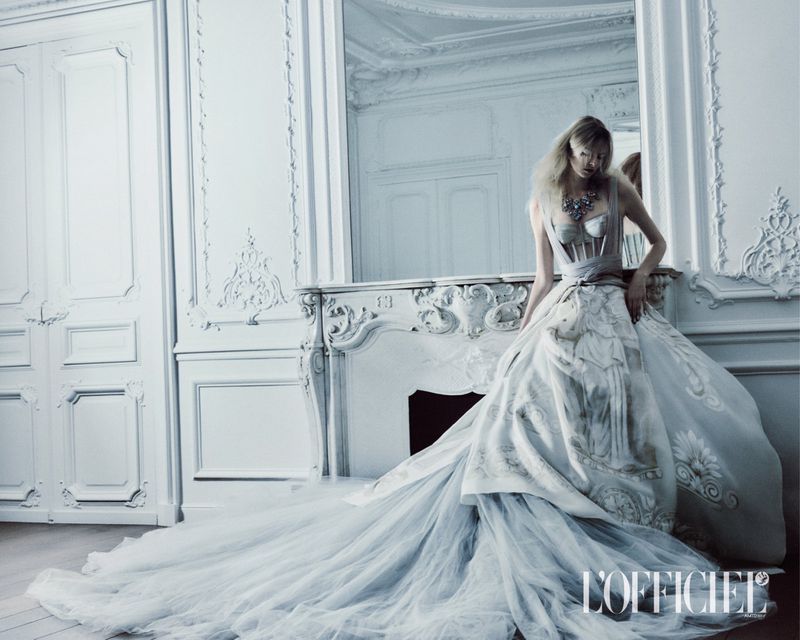 Model in a voluminous white haute couture gown posing against an ornate neoclassical fireplace in an opulent white interior.
