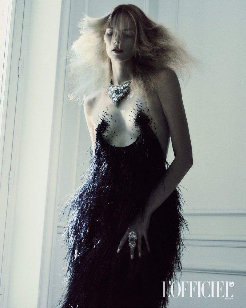 Model Elza Matiz wearing a black feather dress and statement necklace against a white panel wall for L'Officiel Italia editorial
