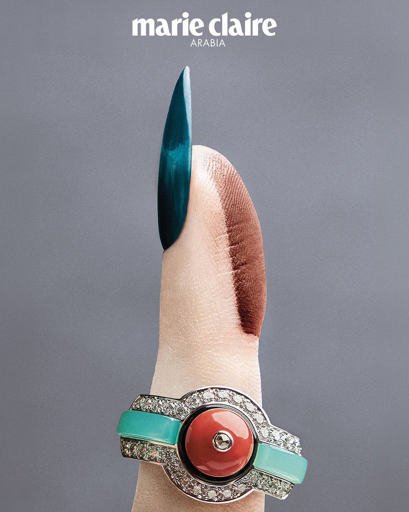 Close-up of a finger with a long teal manicure wearing an ornate diamond and gemstone ring against grey background