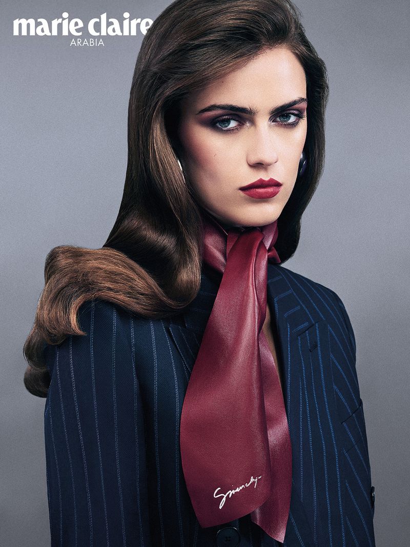 Close-up portrait of a woman wearing a dark pinstripe blazer and a burgundy silk neck scarf against a grey studio backdrop.