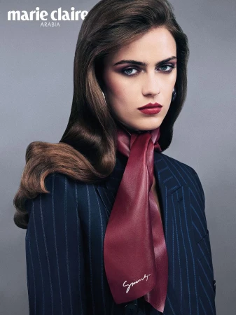 Close-up portrait of a woman wearing a dark pinstripe blazer and a burgundy silk neck scarf against a grey studio backdrop.