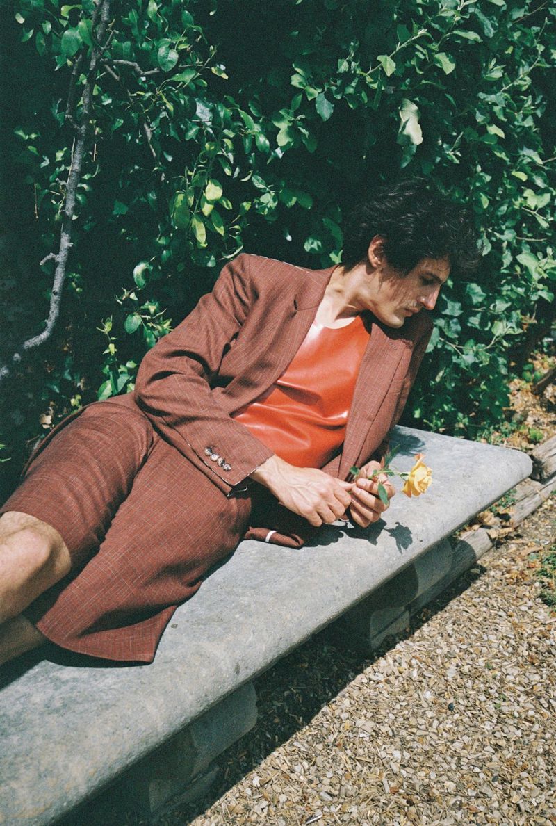 Model lying on a stone bench holding a yellow rose wearing a brown suit and red top against a leafy garden background