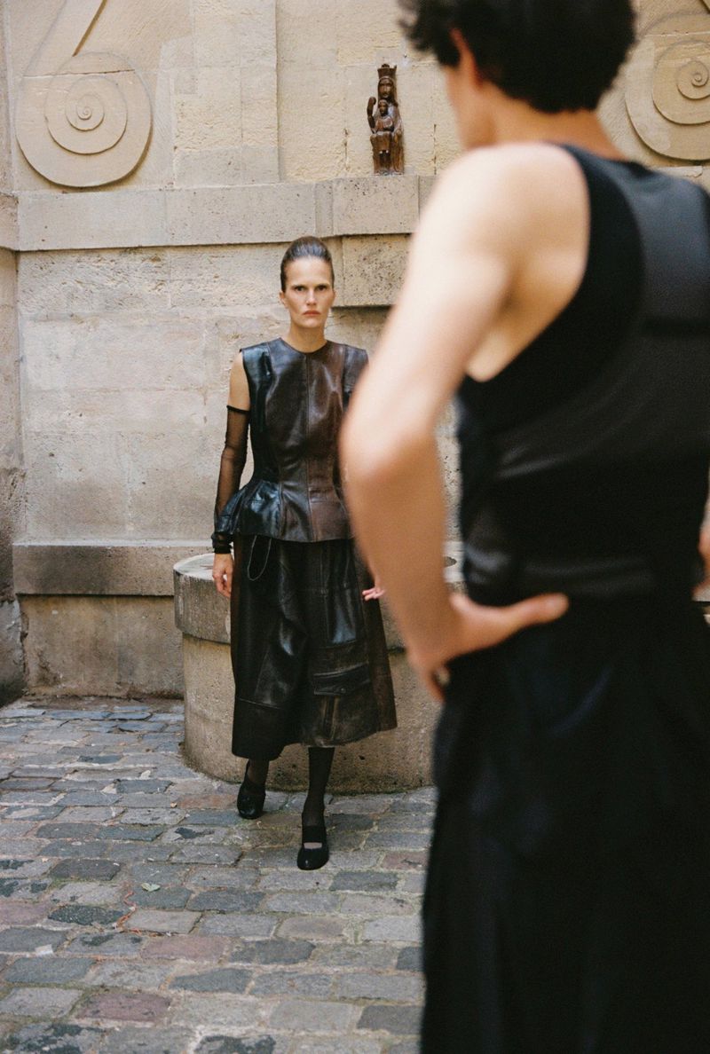 Model standing near a stone well outdoors while a second person is viewed from behind in the foreground