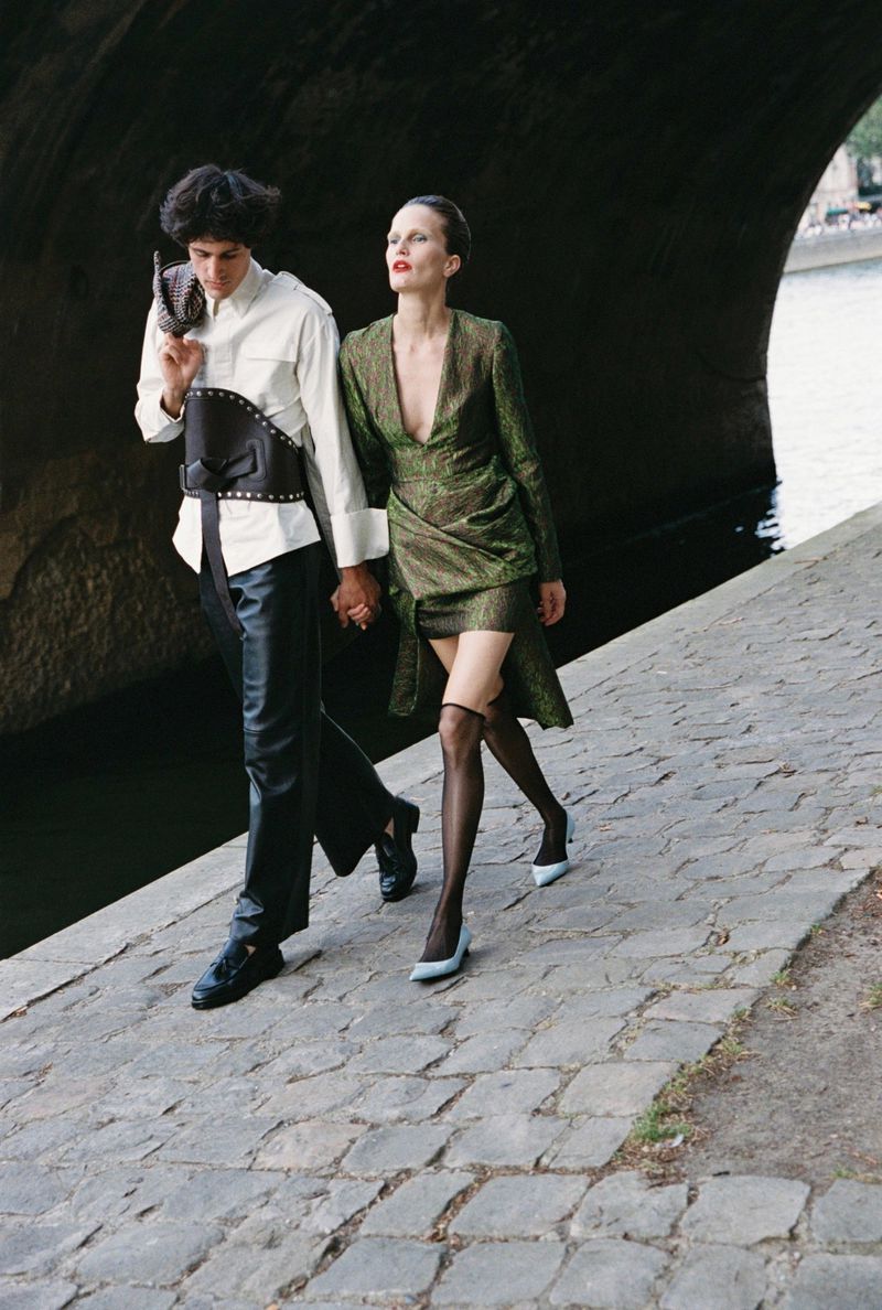 Two models walking together along a stone riverbank under a large bridge for Harper's Bazaar Kazakhstan