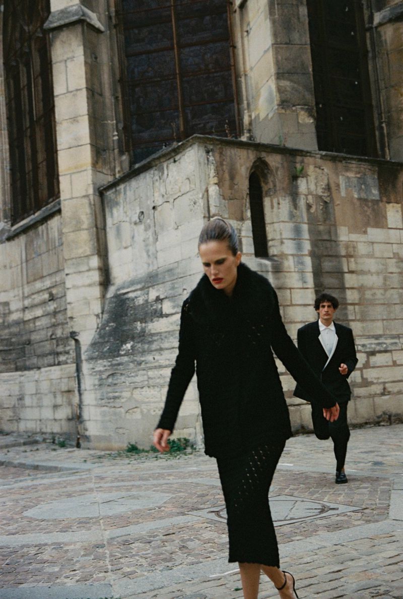Woman in black coat walking outdoors with a man behind her in front of a stone building