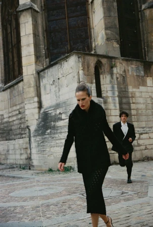 Woman in black coat walking outdoors with a man behind her in front of a stone building