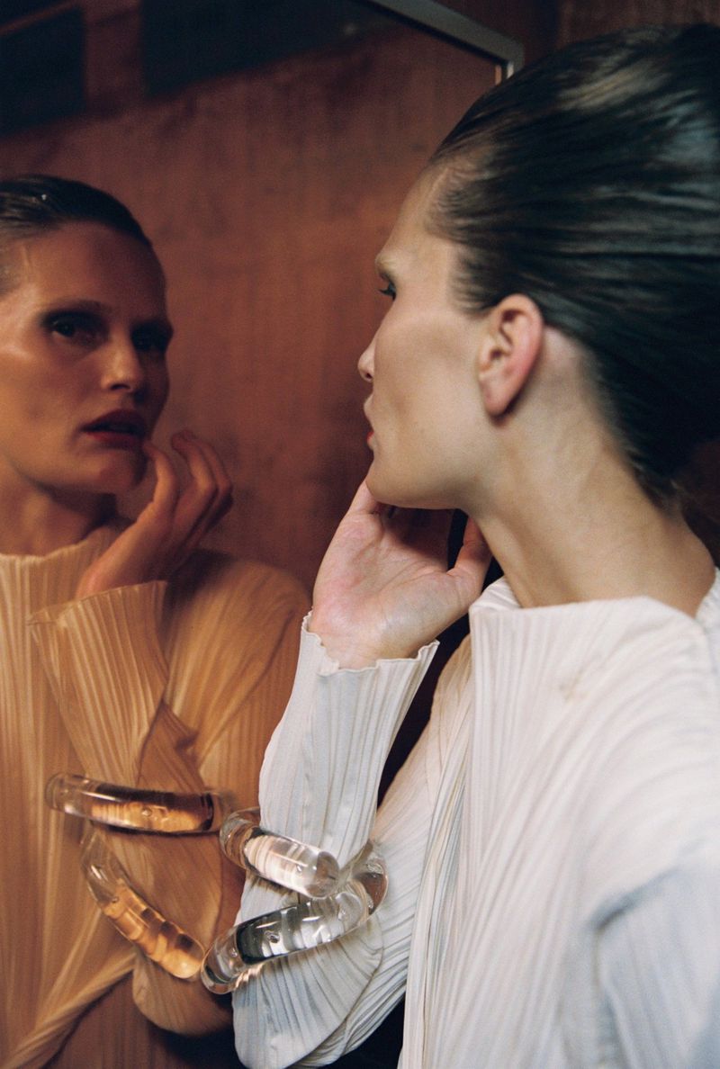 Model touching her reflection in a mirror wearing a pleated white top and clear twisted bracelet