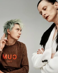 Portrait of two models against a grey background, featuring a young man with mint green hair and an older woman in a white jacket