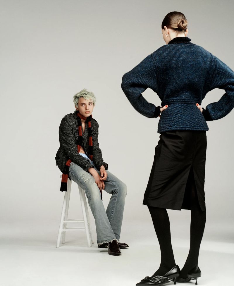 Two models pose in a studio with a grey seamless background, one seated on a white stool wearing a textured jacket and jeans.