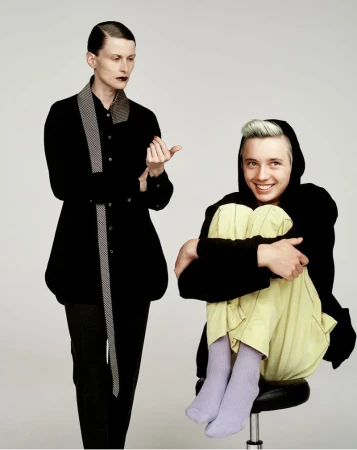 Two models posed against a light grey background for i-D Magazine, featuring a standing woman in black and a seated person hugging their knees.
