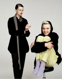 Two models posed against a light grey background for i-D Magazine, featuring a standing woman in black and a seated person hugging their knees.