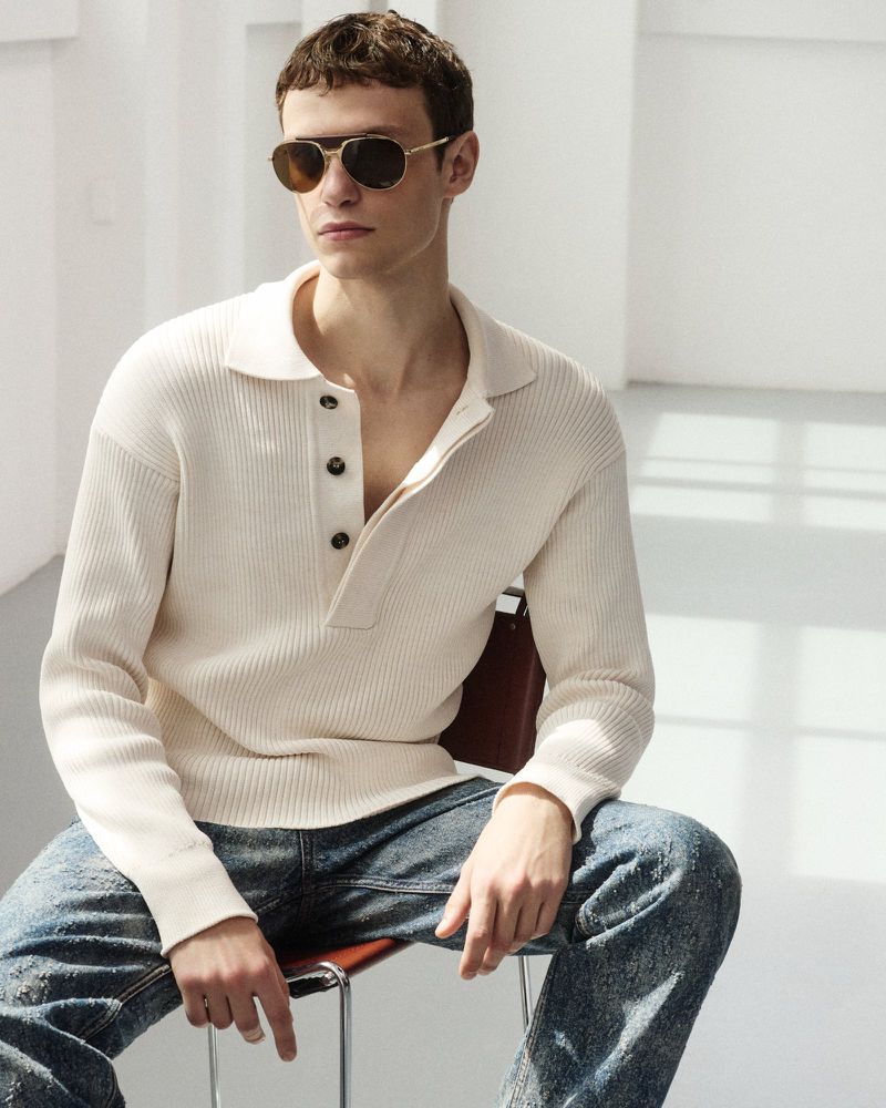 A young adult man with sunglasses sits on a metal chair, wearing a buttoned knit shirt, and blue jeans in a bright indoor setting.