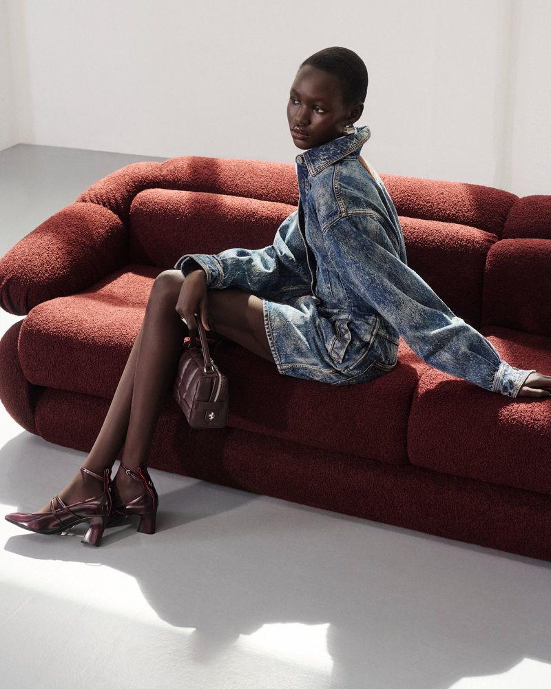 A striking studio shot of a dark-skinned woman in a denim outfit, posing on a textured rust-colored sofa with a matching purse and heels.
