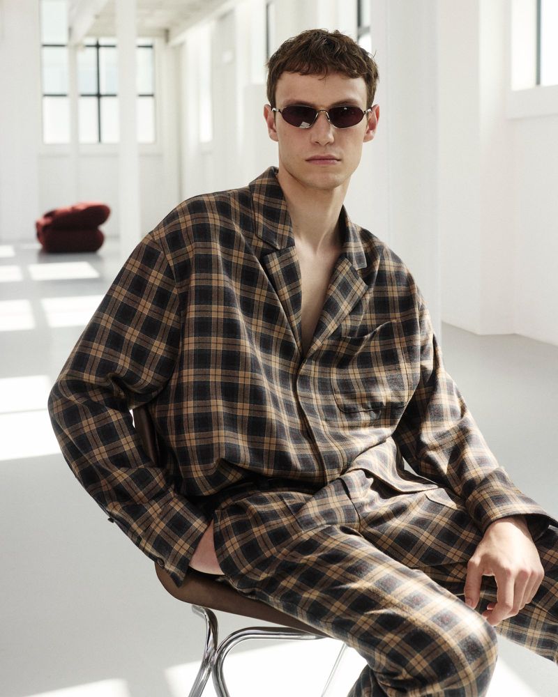 Man sits on a metal stool in a minimalist white room, wearing a tan and black plaid pajama-style outfit and oval sunglasses.