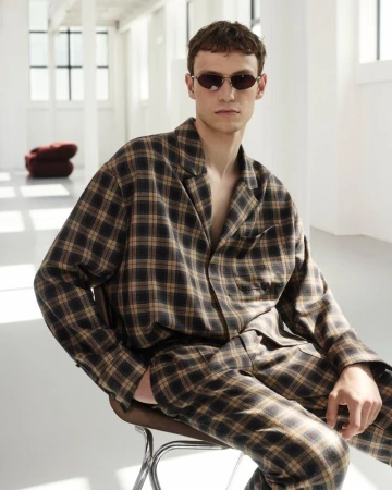 Man sits on a metal stool in a minimalist white room, wearing a tan and black plaid pajama-style outfit and oval sunglasses.