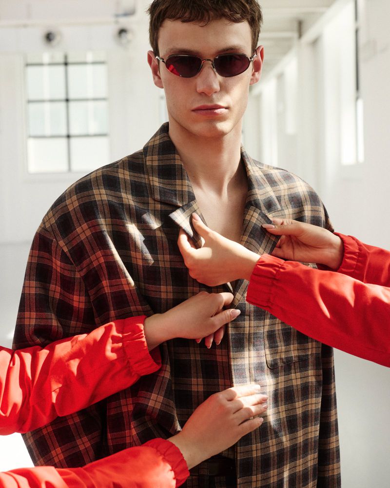 Indoor fashion shot of a slender adult male wearing sunglasses and an oversized plaid jacket, being adjusted by two women in red tops.