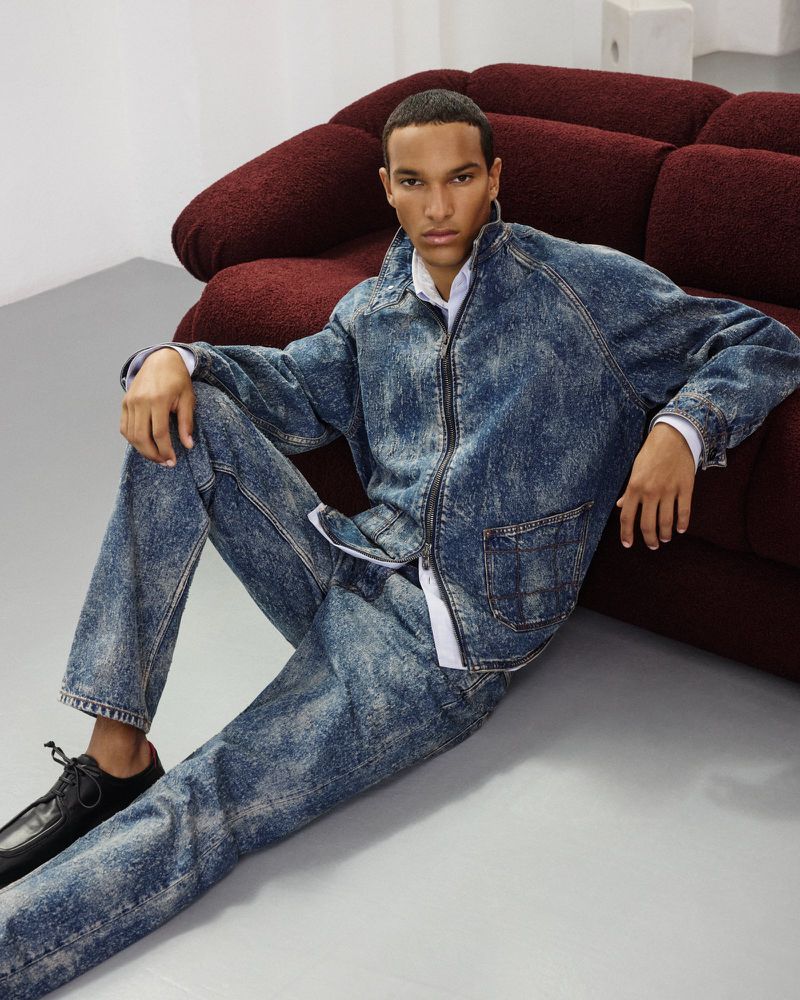 Male model sits near a burgundy sofa wearing a blue acid-wash denim jacket and matching jeans with black leather shoes indoors.