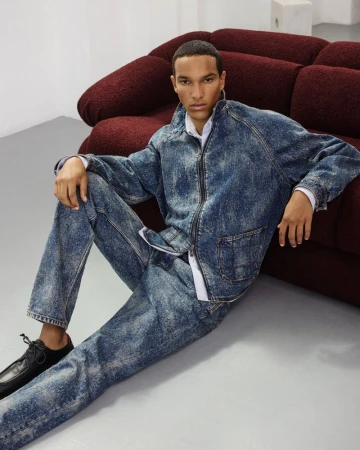 Male model sits near a burgundy sofa wearing a blue acid-wash denim jacket and matching jeans with black leather shoes indoors.