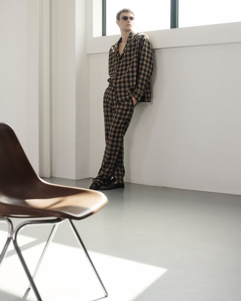 A young adult male wearing a brown checkered pajama set and sunglasses poses leaning against a white wall in front of a window.