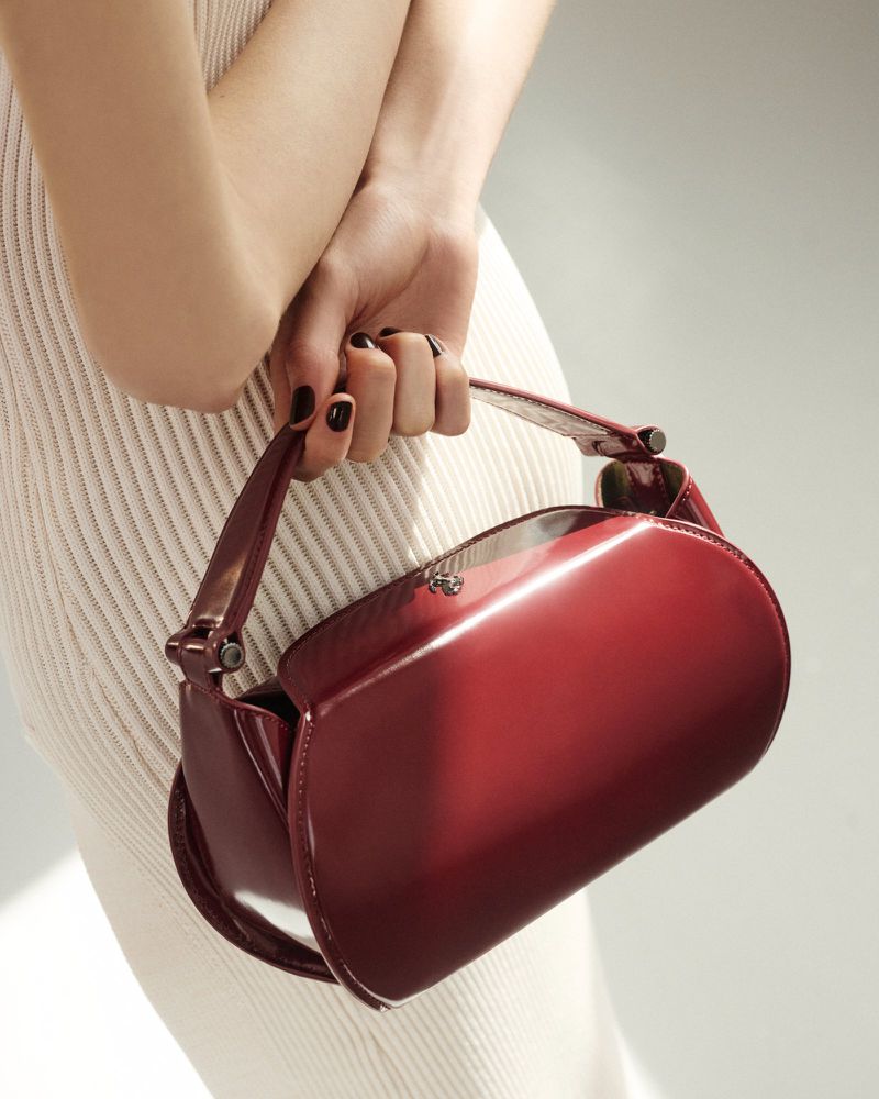 Close-up studio shot of an adult female with dark nail polish holding a shiny, burgundy patent-leather handbag against her body.