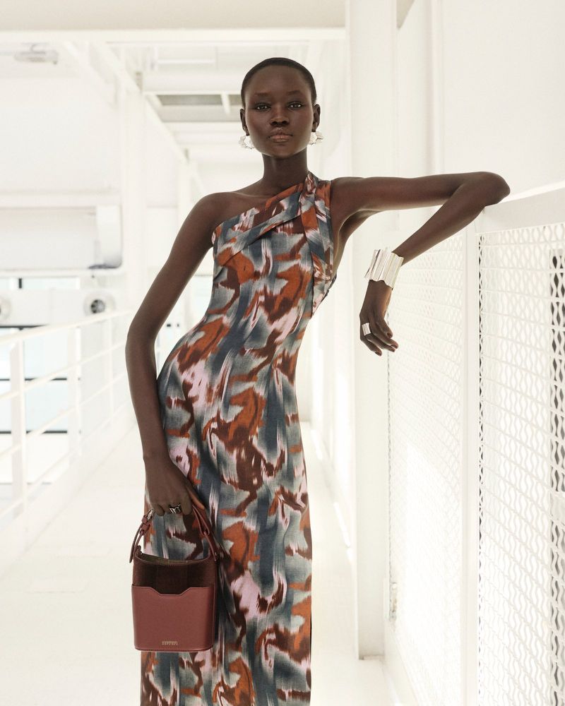 Fashionable adult female posing indoors in a one-shoulder dress holding a purse, complemented by jewelry, against a clean white backdrop.