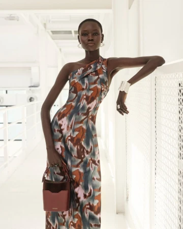 Fashionable adult female posing indoors in a one-shoulder dress holding a purse, complemented by jewelry, against a clean white backdrop.