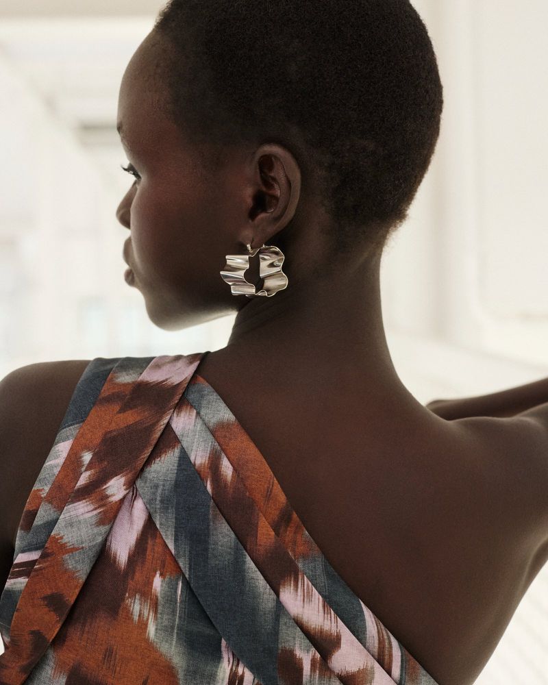 Close-up of a woman with short hair and a patterned dress wearing large, textured silver earrings in a softly lit indoor setting.