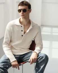 Model wearing a cream ribbed polo shirt and sunglasses sitting on a chair against a white background