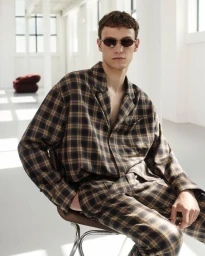 A man wearing a brown plaid shirt and matching trousers seated in a bright white studio space.