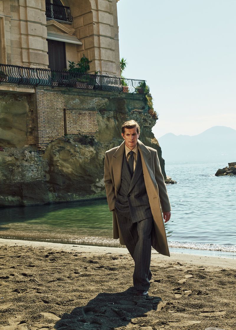 Man wearing a camel coat and grey suit walking on a sandy beach below a classical building