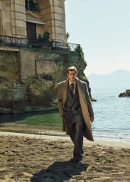 Man wearing a camel coat and grey suit walking on a sandy beach below a classical building