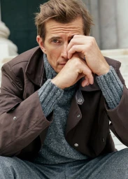 Man wearing a brown jacket over a grey turtleneck sweater with hand raised near his face outdoors.