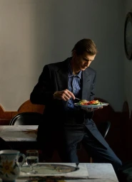 A man in a pinstriped suit jacket eating a salad plate in an indoor setting photographed by Raffo Marone